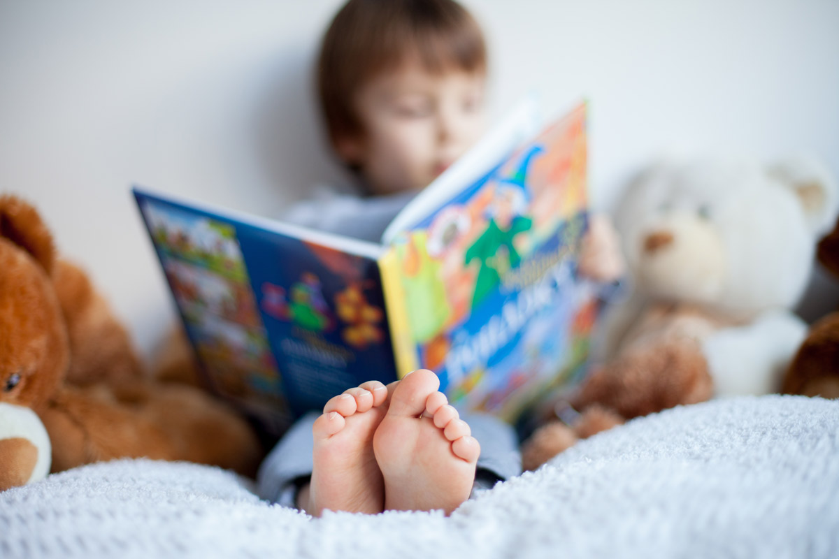 young boy reads a book in bed surrounded by stuffed animals