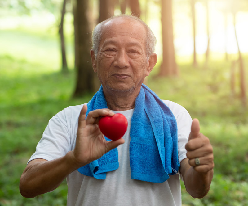 elderly man with a sports towel draped over his shoulders standing outside in a wooded area holding a small felt heart