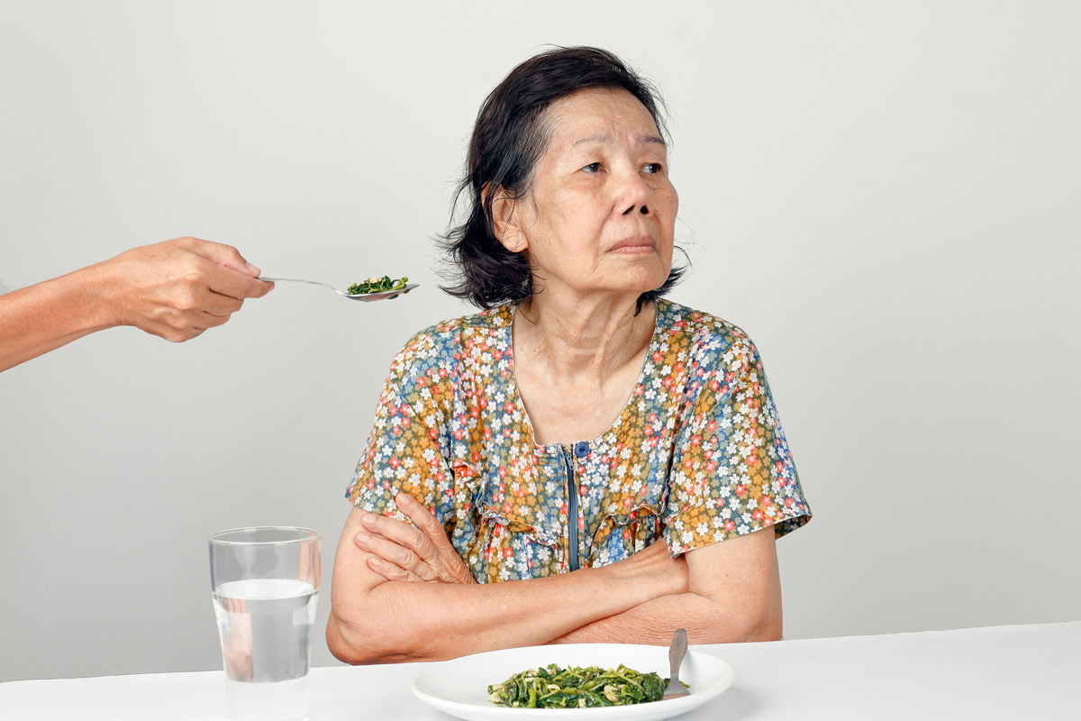 elderly woman with arms crossed refusing to eat a spoonful of food presented to her by a loved one