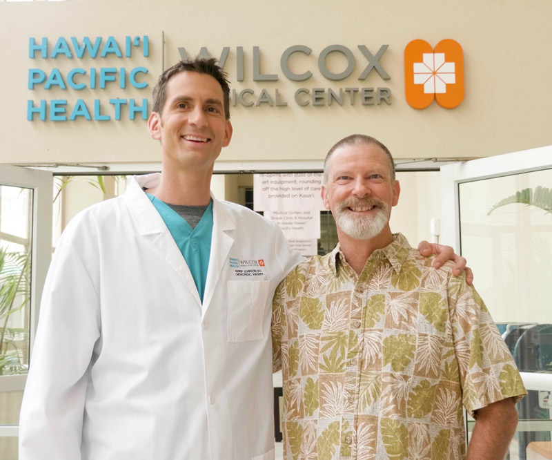 Wilcox orthopedic surgeon Dr. Derek Johnson and Mike Wehrly stand together smiling in front of the entrance to Wilcox Medical Center