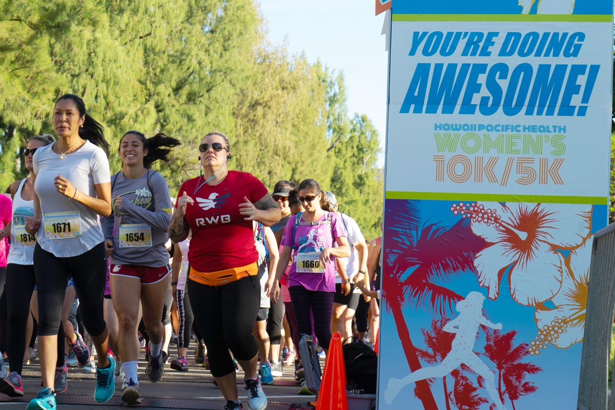 Women cross the starting line of the 2018 Hawaii Pacific Health Women's 10K with the encouraging words of "You're Doing Awesome" printing on the starting arch.