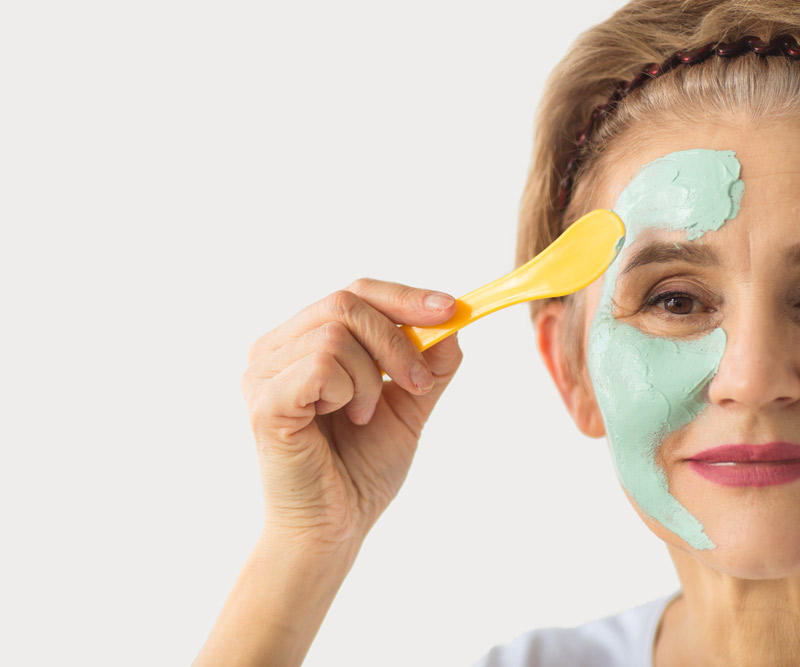 an older woman applies a green face mask to her skin