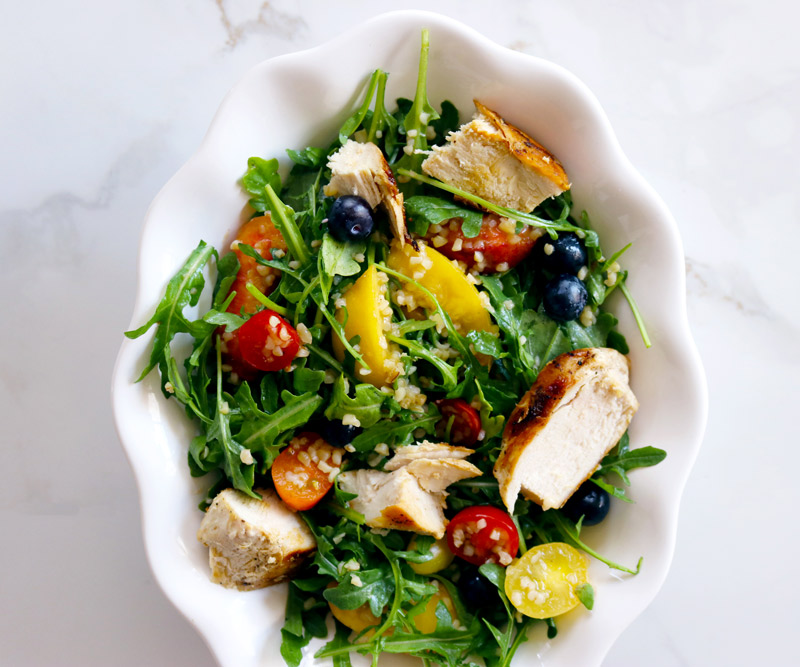 a white bowl filled with Chicken & Bulgur Salad made with arugula, grilled chicken, bulgur, cherry tomatoes, peaches and blueberries sits on a white marble counter top