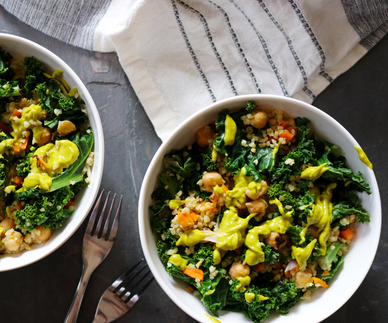 two Kale & Chickpea Harvest Bowls drizzled with bright-green Avocado Dressing arranged on a slate counter top next to two forks and a beige-and-black dish towel