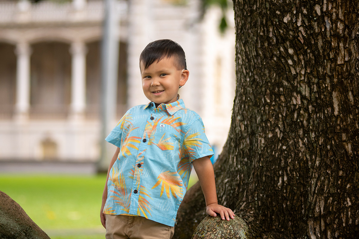 A young boy with a cochlear ear implant wears an aloha shirt and leans against the trunk of a tree in front of Iolani Palace.