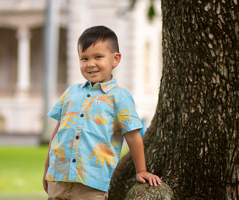 A young boy with a cochlear ear implant wears an aloha shirt and leans against the trunk of a tree in front of Iolani Palace.