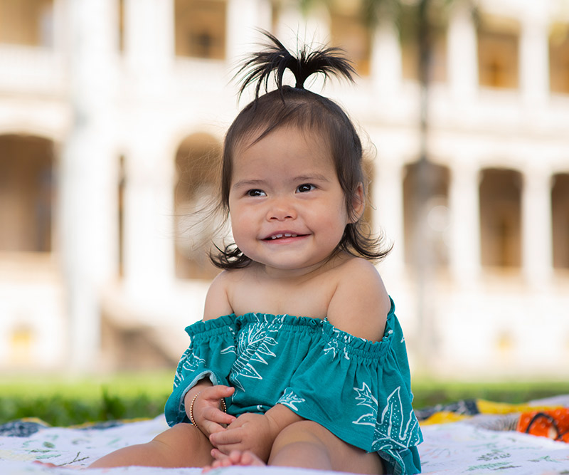 A 9-month-old girl sits on a colorful blanket in front of Iolani Palace and smiles at the camera.