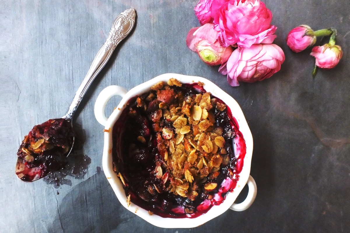 a white ramekin of Old-Fashioned Fruit Crumble with a spoonful of crumble taken out of it sits on a slate counter top that has been decorated with a bunch of pink flowers