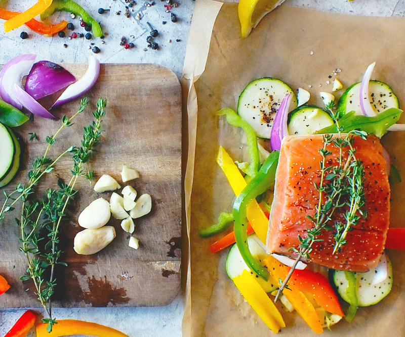 bright orange salmon arranged on a sheet of parchment paper, surrounded by sliced zucchini, bell peppers and onions next to a cutting board with more freshly sliced veggies, garlic and herbs
