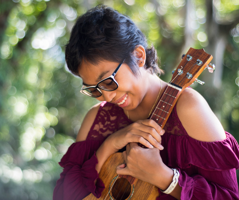 A young girl with short brown hair and glasses smiles while holding an ukulele.