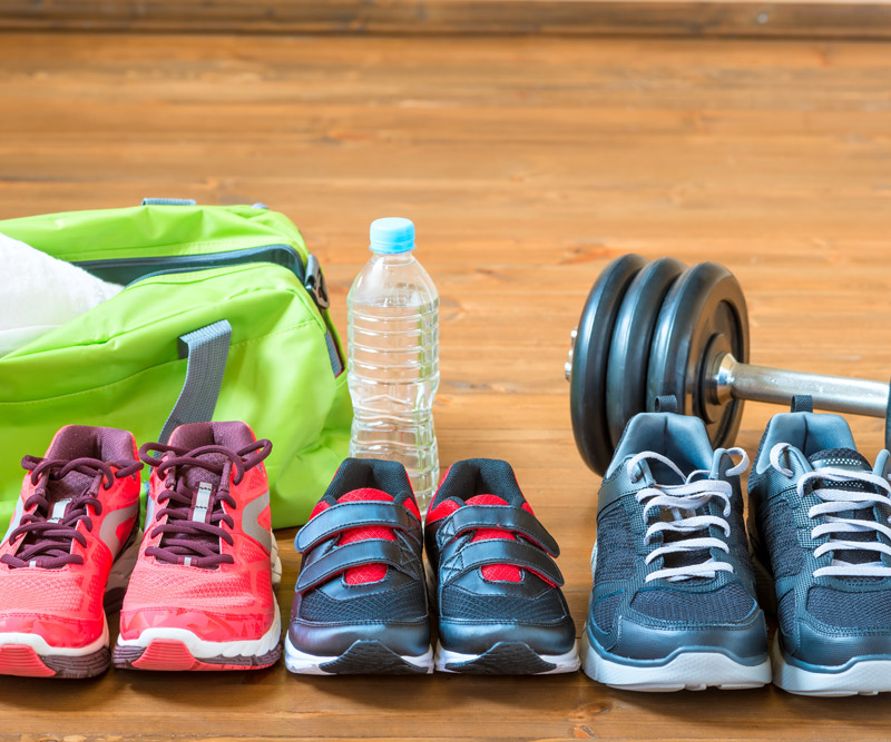 A pair of woman's, child's and man's running shoes sit in front of a gym bag, water bottle and weights to represent a family getting into shape together.