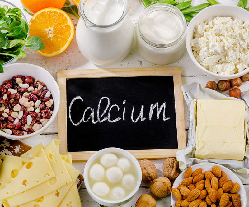 Calcium-rich foods, including milk, cheese, nuts, and leafy greens, displayed on a table around a handheld chalkboard with the word "Calcium" written on it