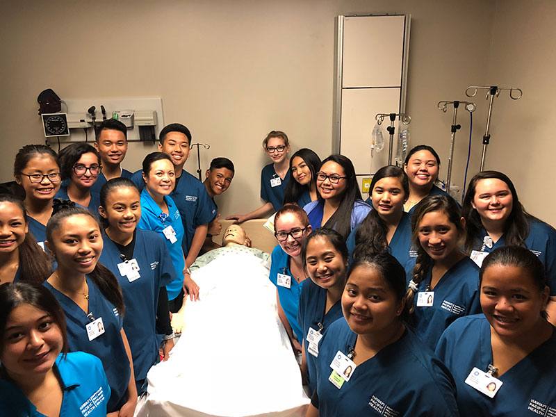 Group of students in medical scrubs gathered around a hospital bed with simulation patient manikin.