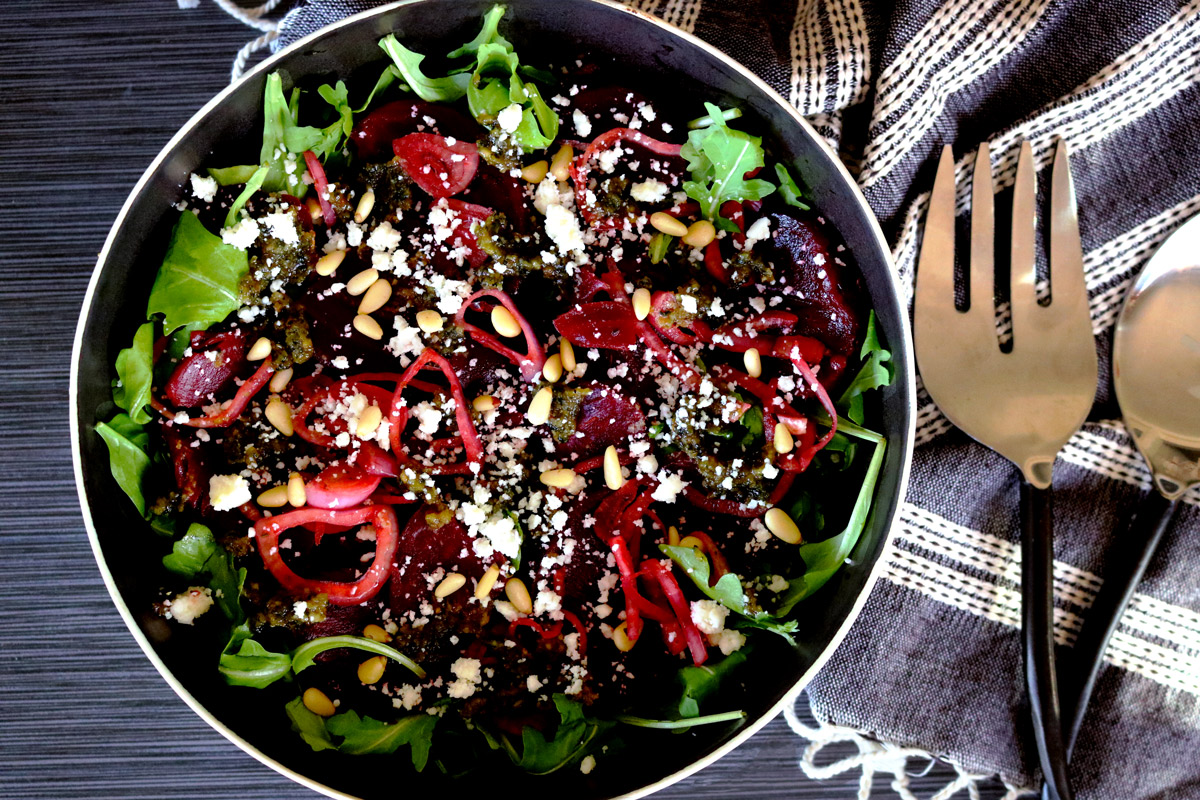 a bowl of Roasted Beet Salad with Basil-Lemon Pesto sits on a dark counter top next to a napkin and serving utensils