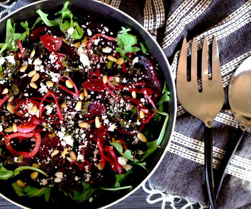 a bowl of Roasted Beet Salad with Basil-Lemon Pesto sits on a dark counter top next to a napkin and serving utensils