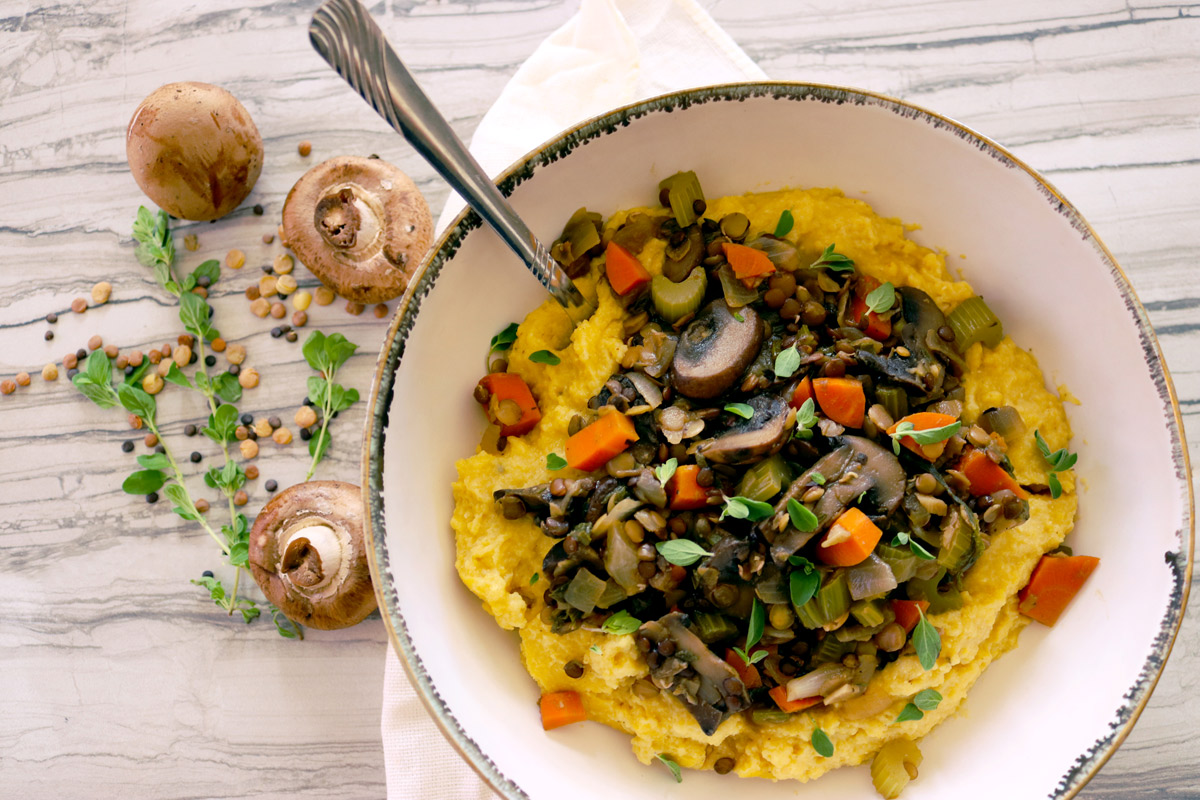 a big bowl of Lentil & Mushroom Stew with Creamy Polenta sits on a wooden table set with a napkin, with loose mushrooms, dry lentils and herbs positioned to the side