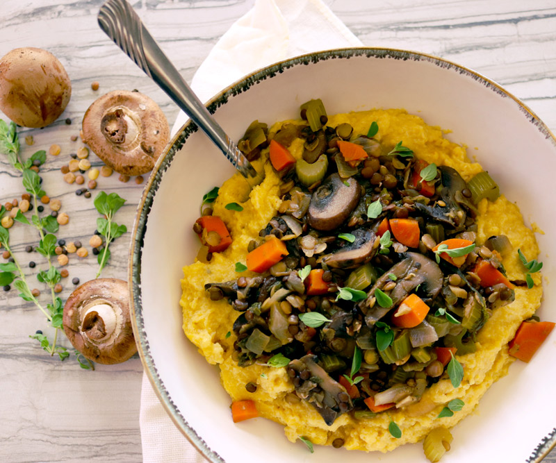 a big bowl of Lentil & Mushroom Stew with Creamy Polenta sits on a wooden table set with a napkin, with loose mushrooms, dry lentils and herbs positioned to the side