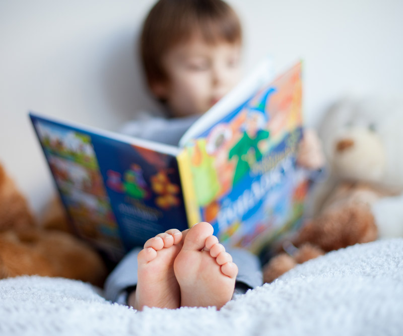 young boy reads a book in bed surrounded by stuffed animals