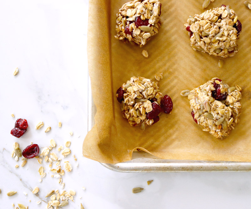 4-Ingredient Breakfast Cookies on a parchment-lined baking sheet sit on a marble counter top sprinkled with oats, sunflower seed kernels and dried cranberries