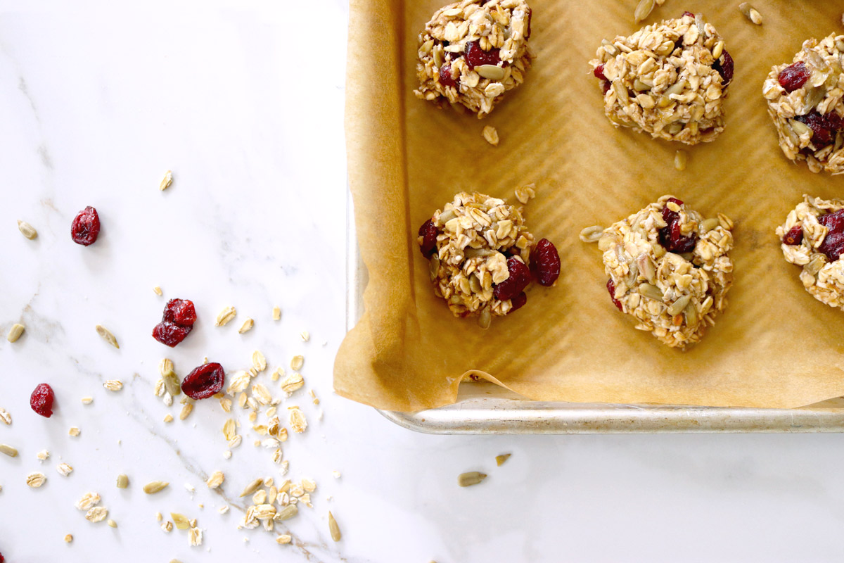 4-Ingredient Breakfast Cookies on a parchment-lined baking sheet sit on a marble counter top sprinkled with oats, sunflower seed kernels and dried cranberries