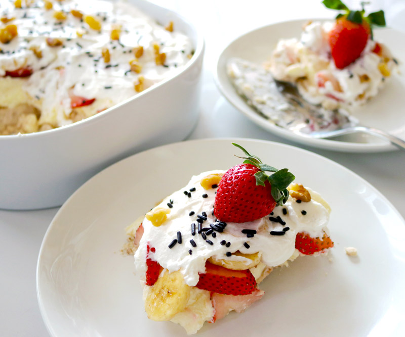 two slices of No-Bake Banana Split Cheesecake topped with fresh strawberries and chocolate sprinkles presented on clean white plates next to a large ceramic baking dish of cheesecake with a large portion sliced out