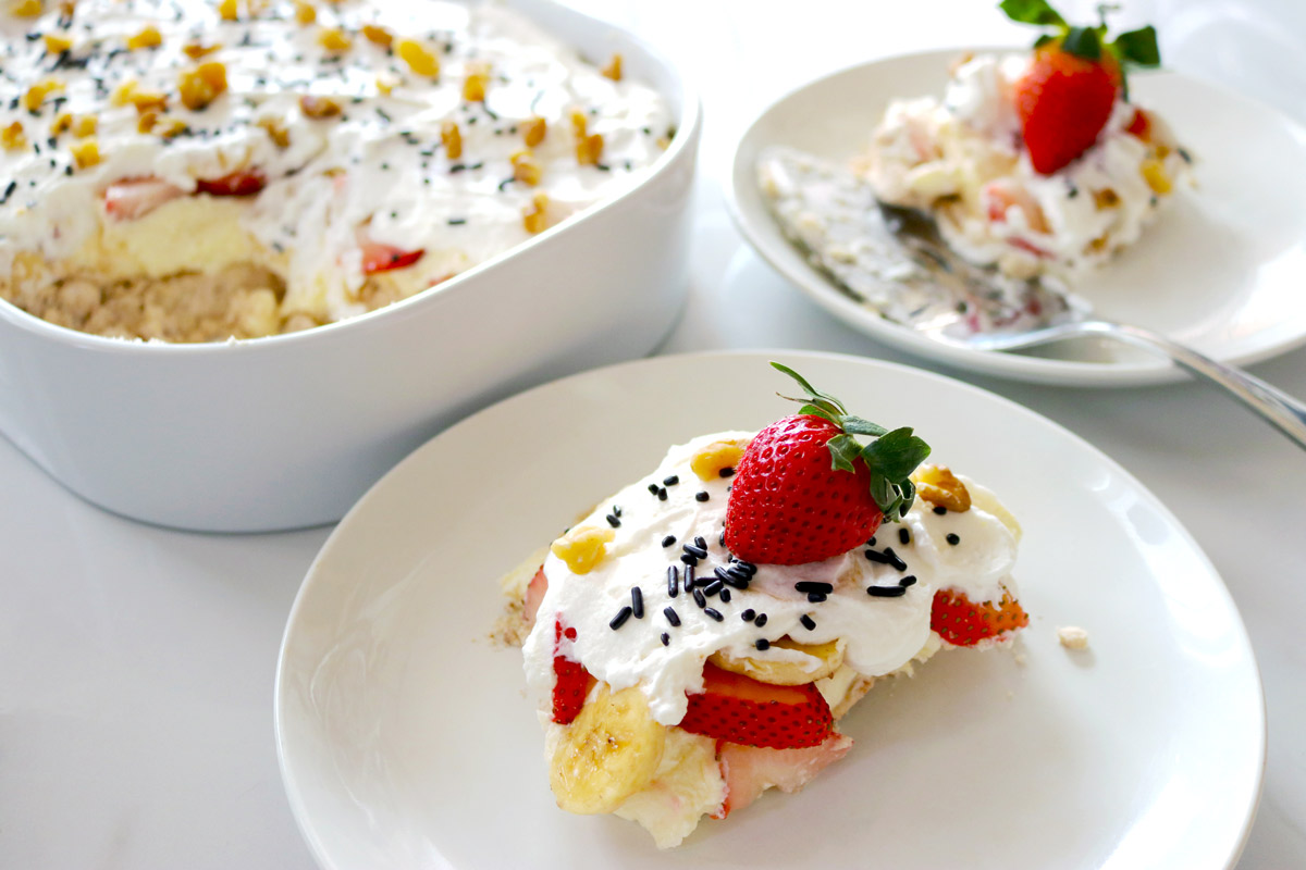 two slices of No-Bake Banana Split Cheesecake topped with fresh strawberries and chocolate sprinkles presented on clean white plates next to a large ceramic baking dish of cheesecake with a large portion sliced out