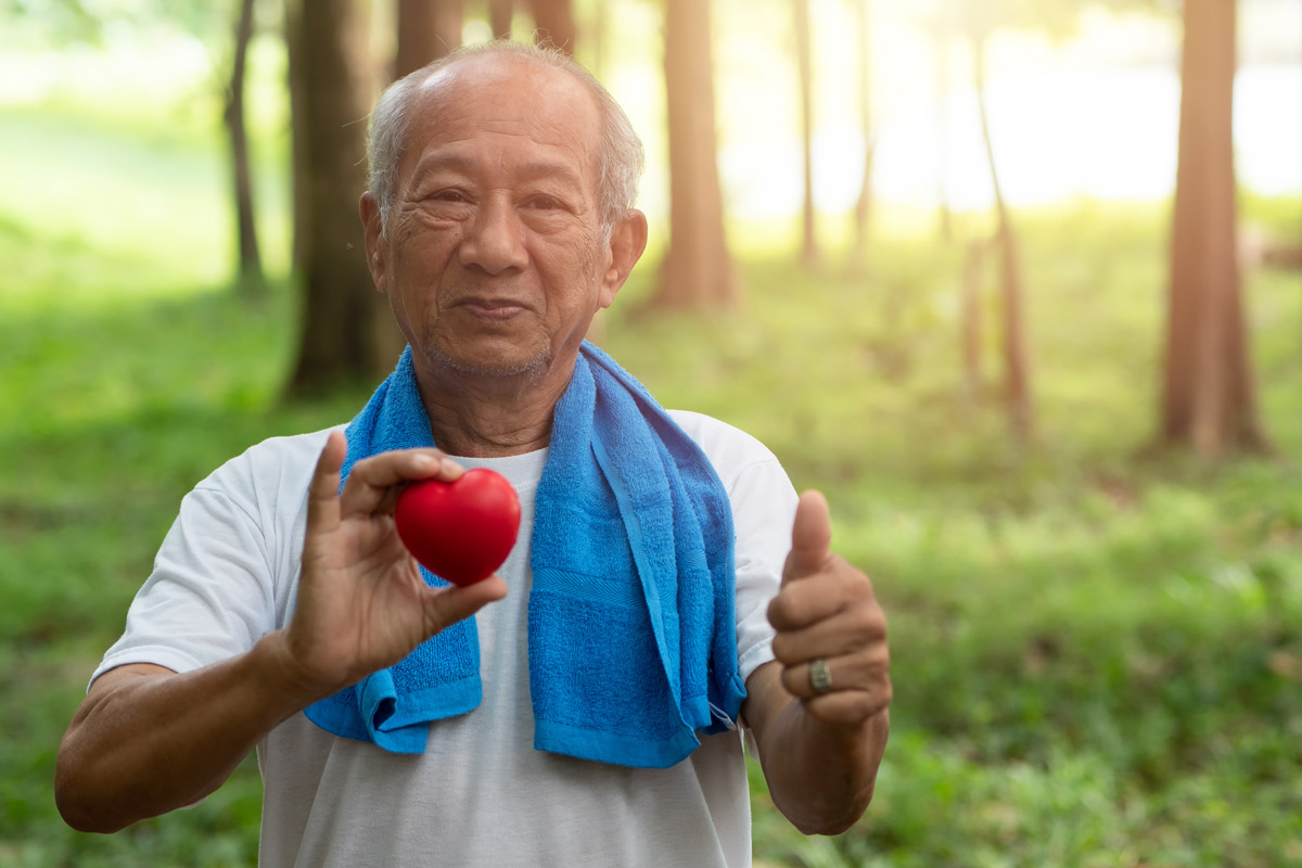 elderly man with a sports towel draped over his shoulders standing outside in a wooded area holding a small felt heart