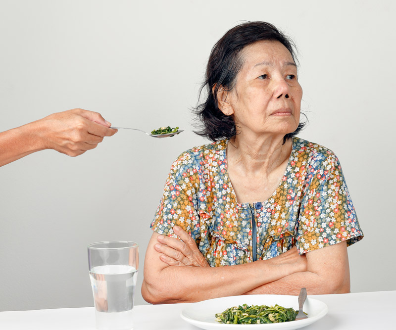 elderly woman with arms crossed refusing to eat a spoonful of food presented to her by a loved one