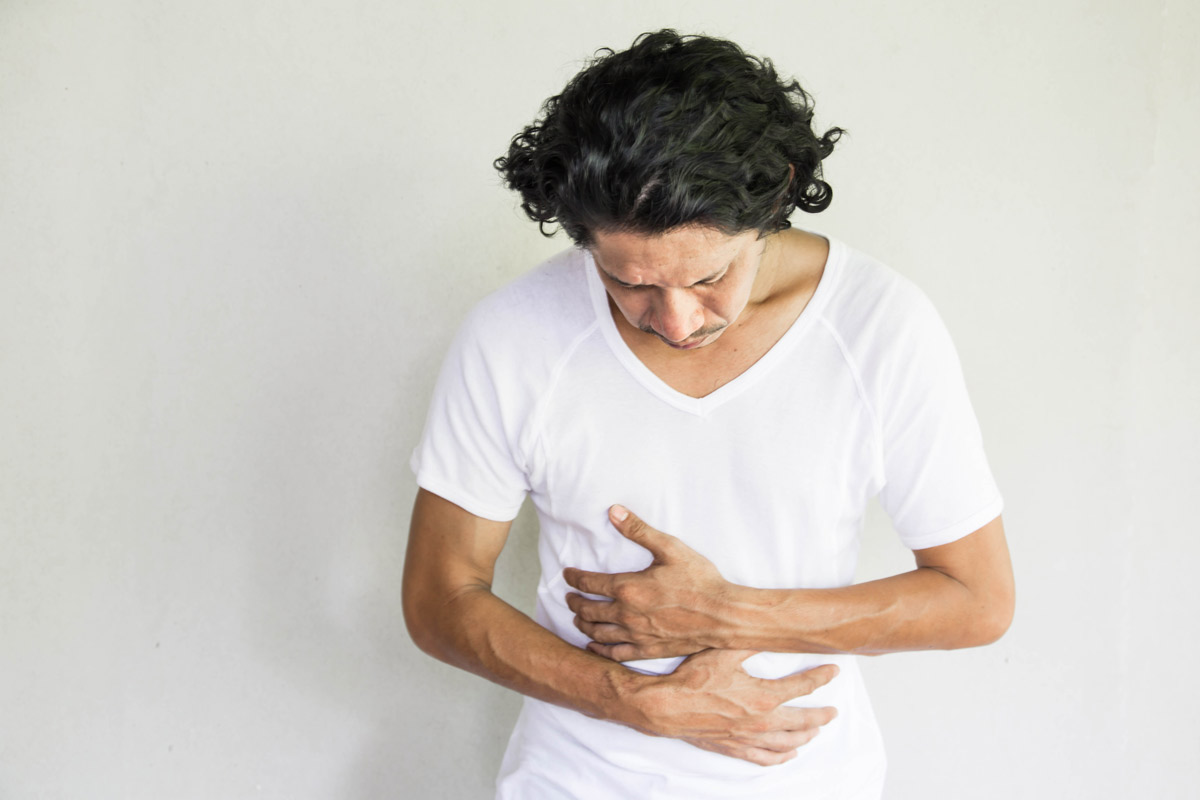 middle-aged man in a white T-shirt holds his stomach with a look of pain on his face to indicate possible colon cancer