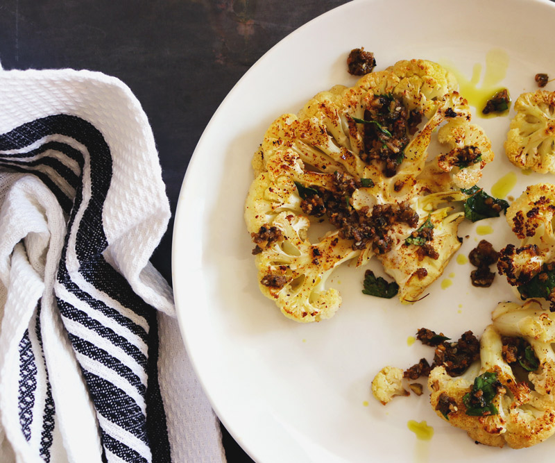 a plate of roasted Cauliflower Steaks drizzled with Spicy Chimichurri Sauce and a white-and-blue-striped napkin sit on a slate counter top