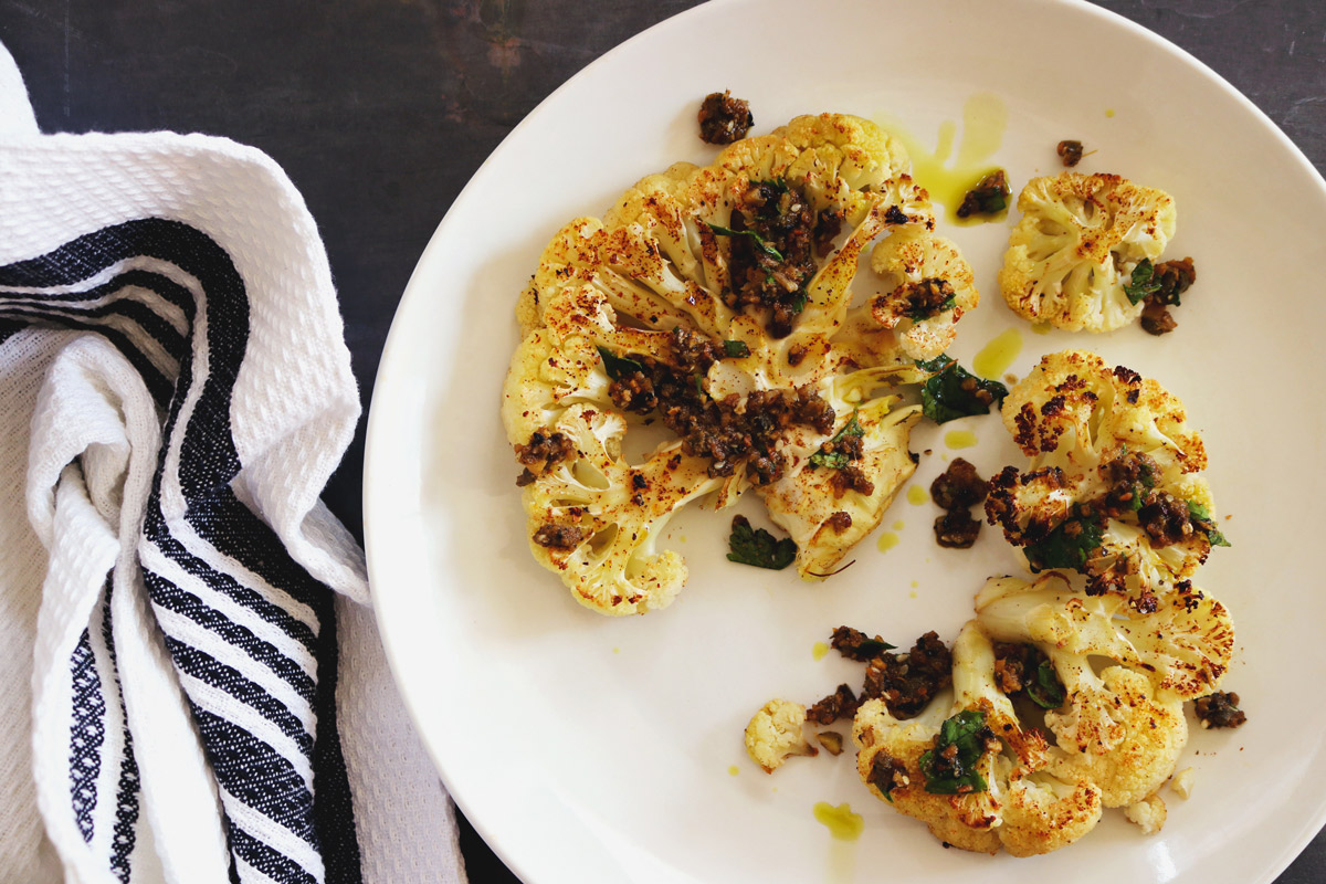 a plate of roasted Cauliflower Steaks drizzled with Spicy Chimichurri Sauce and a white-and-blue-striped napkin sit on a slate counter top