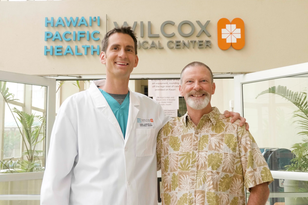 Wilcox orthopedic surgeon Dr. Derek Johnson and Mike Wehrly stand together smiling in front of the entrance to Wilcox Medical Center