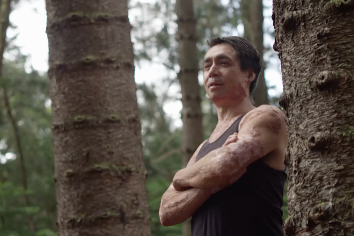 Lance Kaanoi stands in a forest of trees with his arms crossed, looking with determination off into the distance