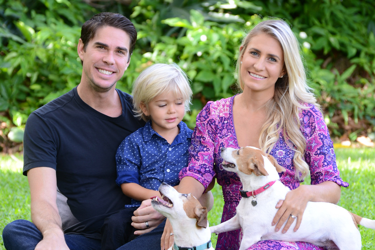 A husband, wife and their young son sit in the grass with two Jack Russell terriers