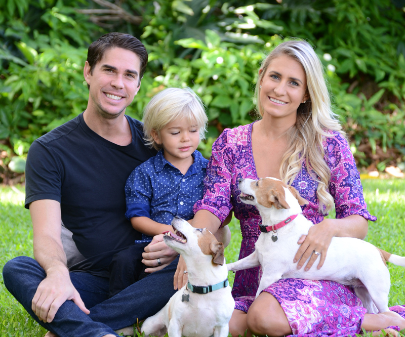 A husband, wife and their young son sit in the grass with two Jack Russell terriers