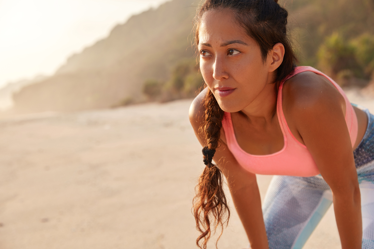 a young woman dressed in running clothes stands with her hands on her knees on a beach