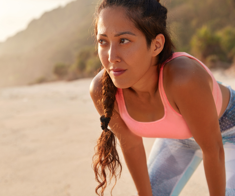 a young woman dressed in running clothes stands with her hands on her knees on a beach