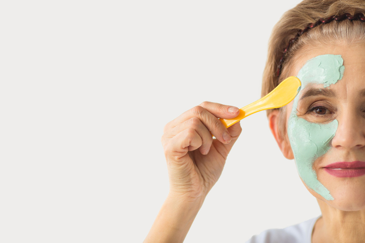 an older woman applies a green face mask to her skin