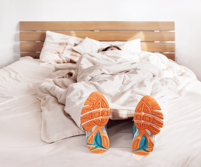 orange-soled athletic shoes stick out from under the covers of a comforter on a bed