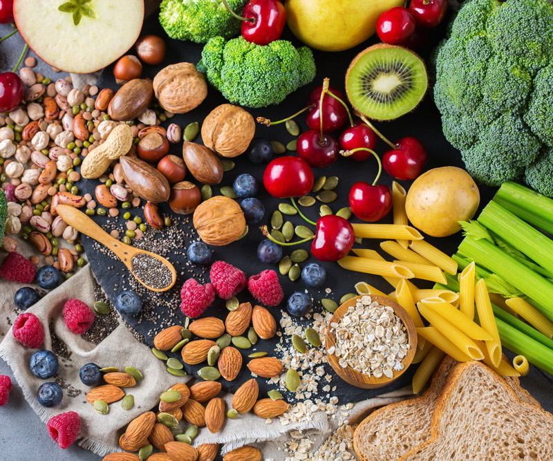 a variety of carb sources, including broccoli, berries, almonds, pumpkin seeds, chia seeds, cherries, apples, oatmeal, bread, legumes and pasta arranged on a slate counter