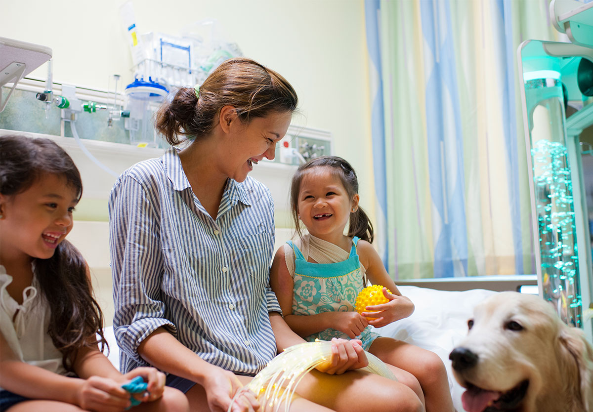 two children and an adult sitting on a bed in a hospital with a dog near them