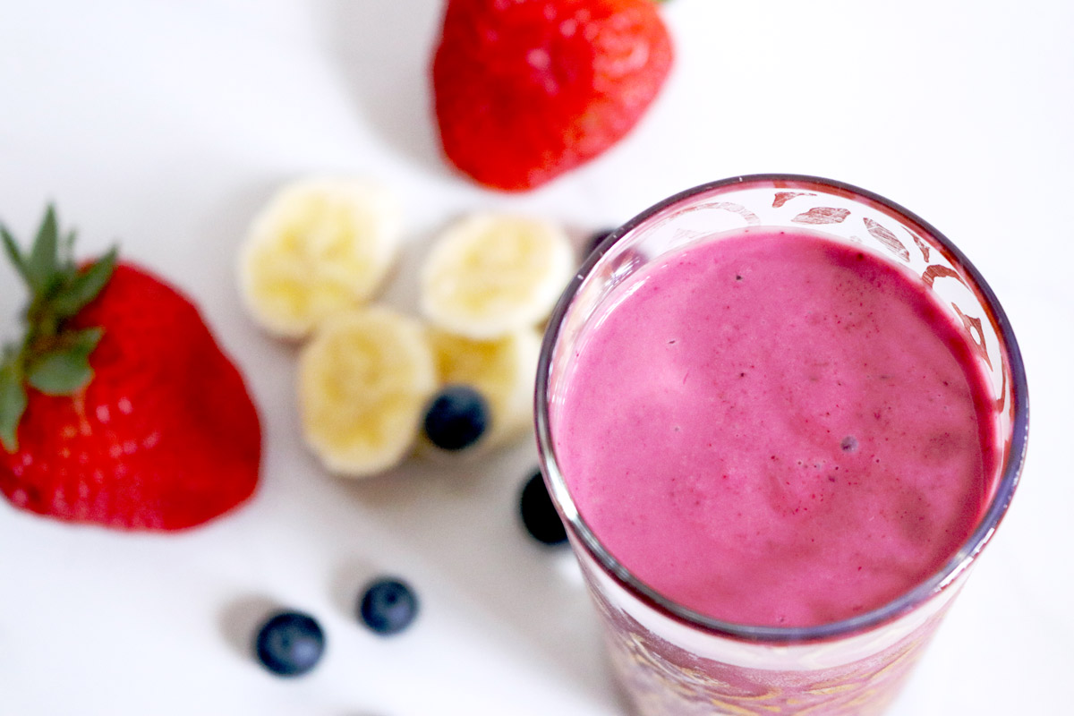 a glass full of bright-pink Mixed Berry Smoothie on a white marble counter top surrounded by strawberries, blueberries and sliced banana
