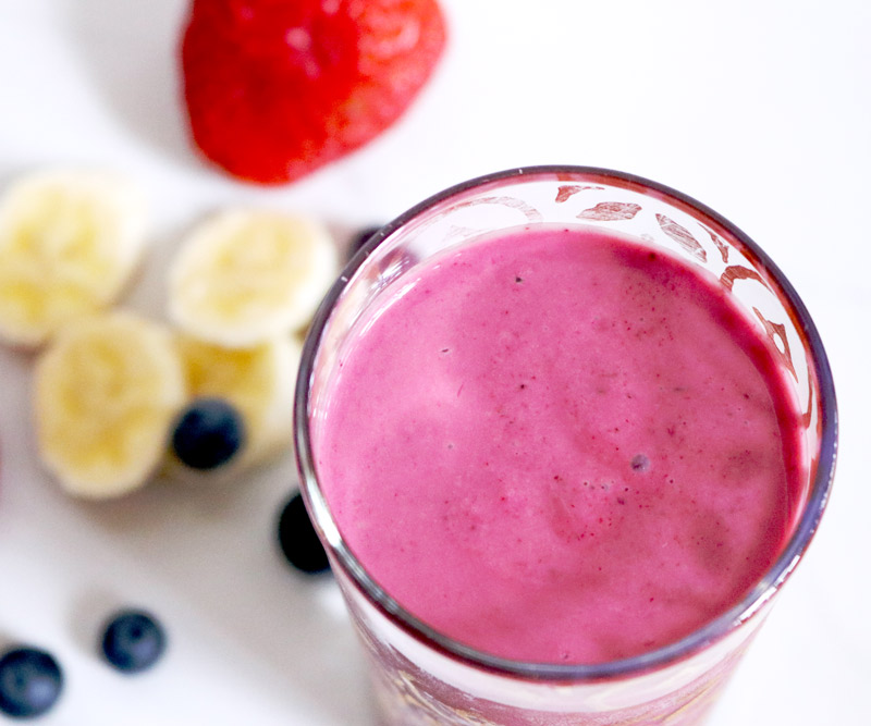 a glass full of bright-pink Mixed Berry Smoothie on a white marble counter top surrounded by strawberries, blueberries and sliced banana