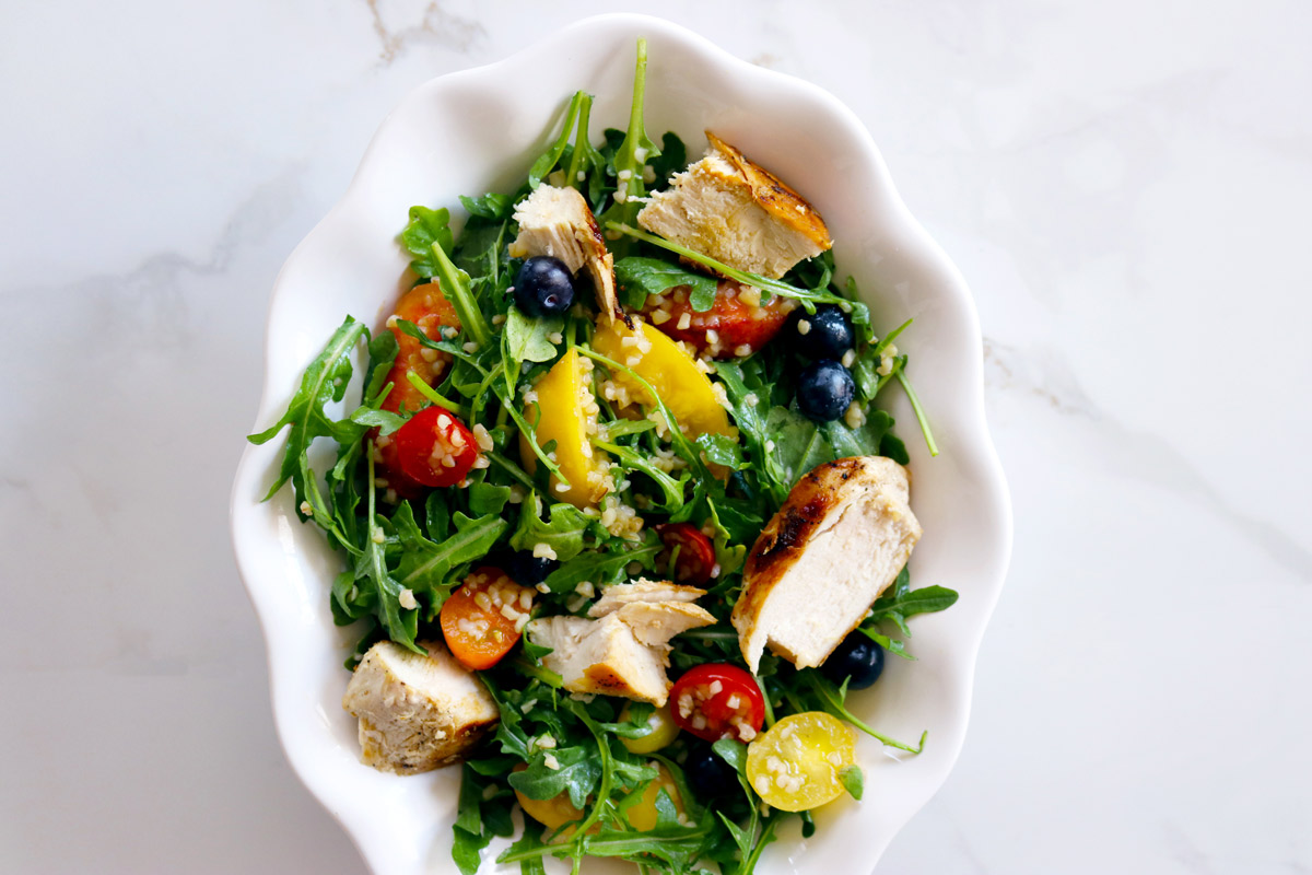 a white bowl filled with Chicken & Bulgur Salad made with arugula, grilled chicken, bulgur, cherry tomatoes, peaches and blueberries sits on a white marble counter top