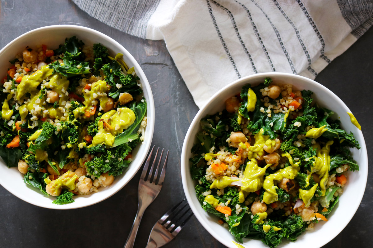 two Kale & Chickpea Harvest Bowls drizzled with bright-green Avocado Dressing arranged on a slate counter top next to two forks and a beige-and-black dish towel