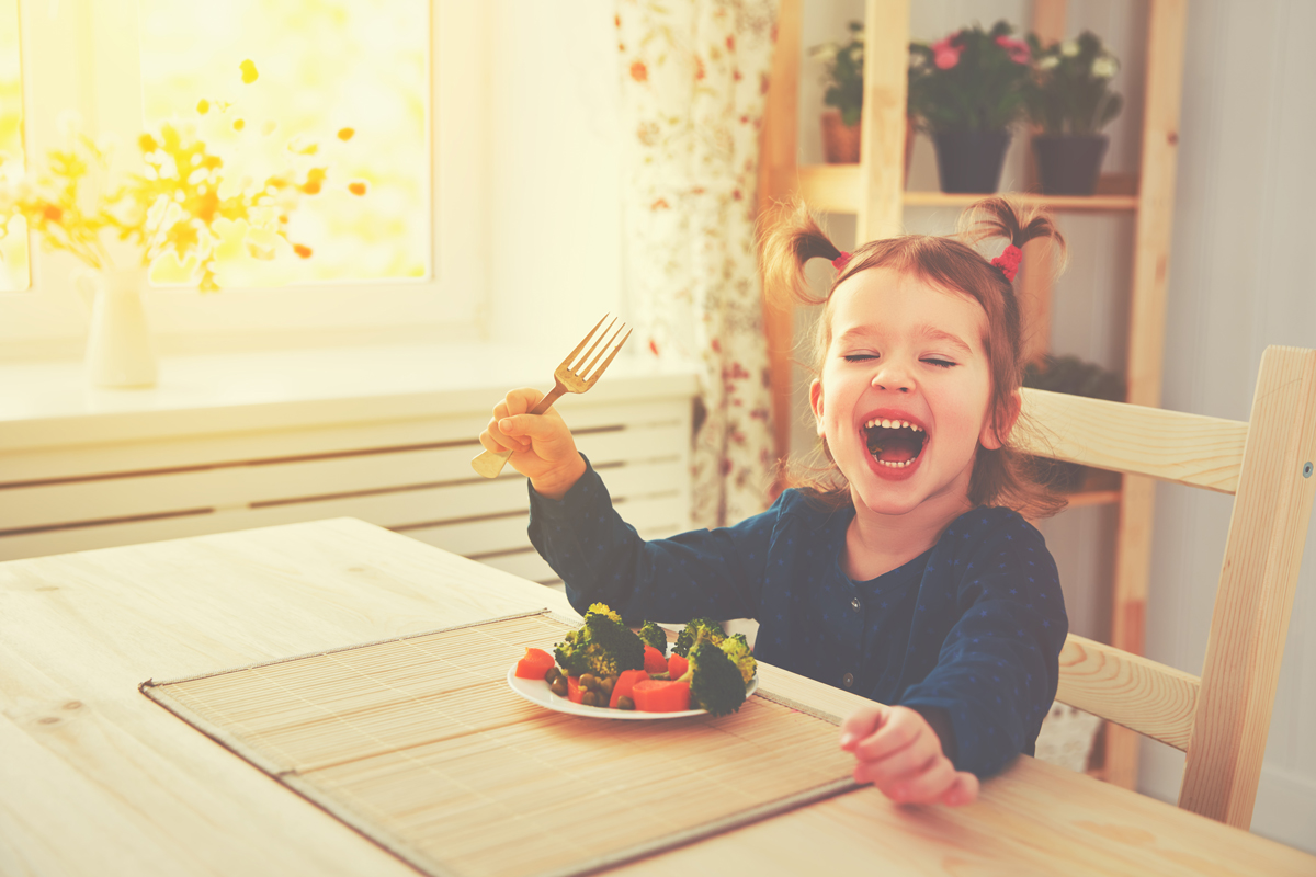 young girl with pigtails sits at a kitchen table with a plateful of vegetables in front of her