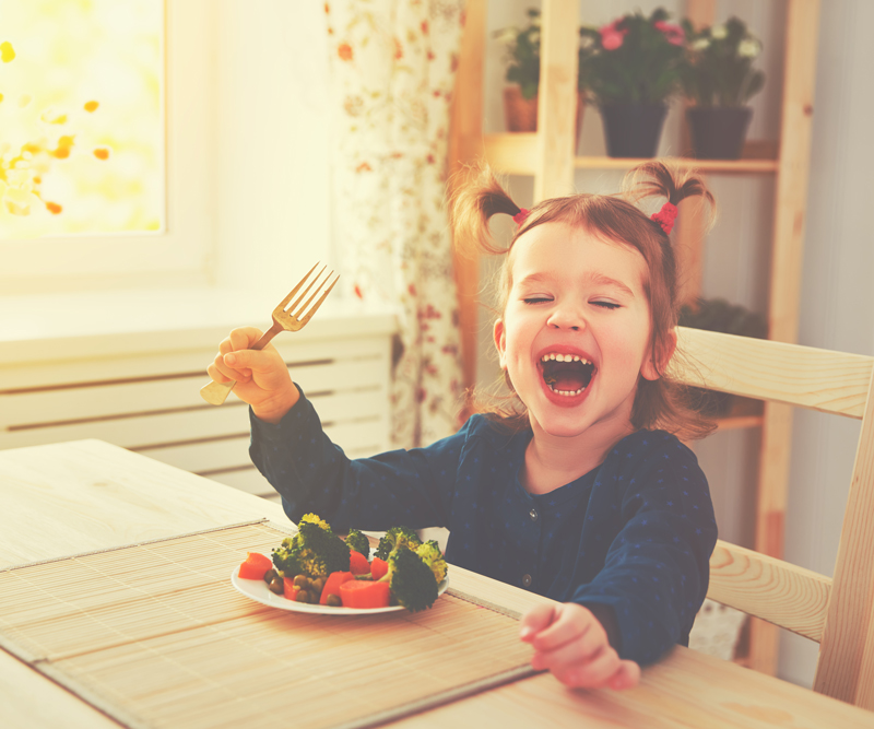 young girl with pigtails sits at a kitchen table with a plateful of vegetables in front of her