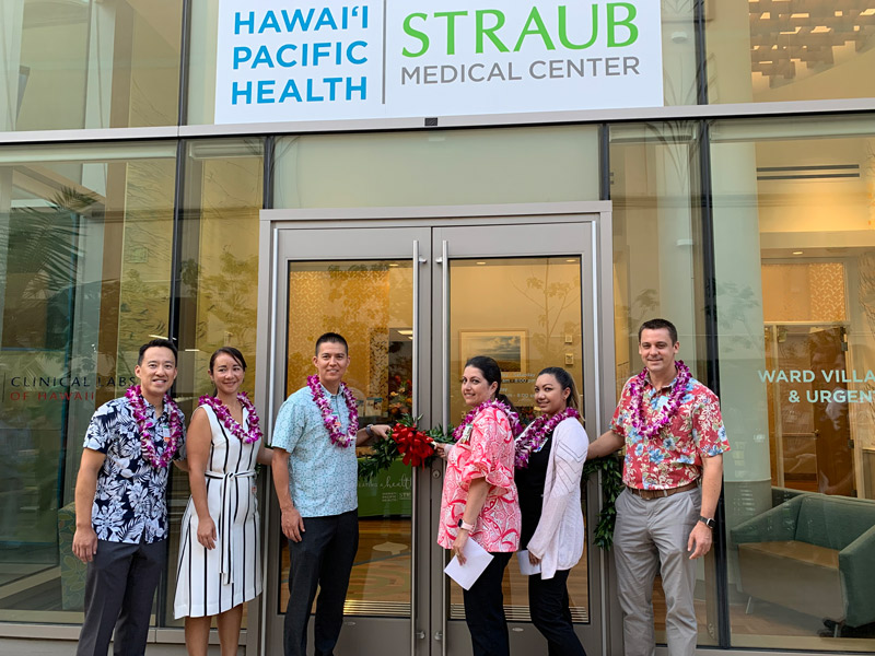 Three men and three women get ready to untie a maile lei draped in front of the glass doors of the new Straub Ward Village clinic