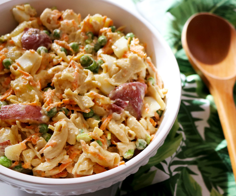 a bowl of Healthier Hawaiian-Style Potato-Mac Salad in a porcelain salad bowl next to a palm-print napkin and wooden serving spoon