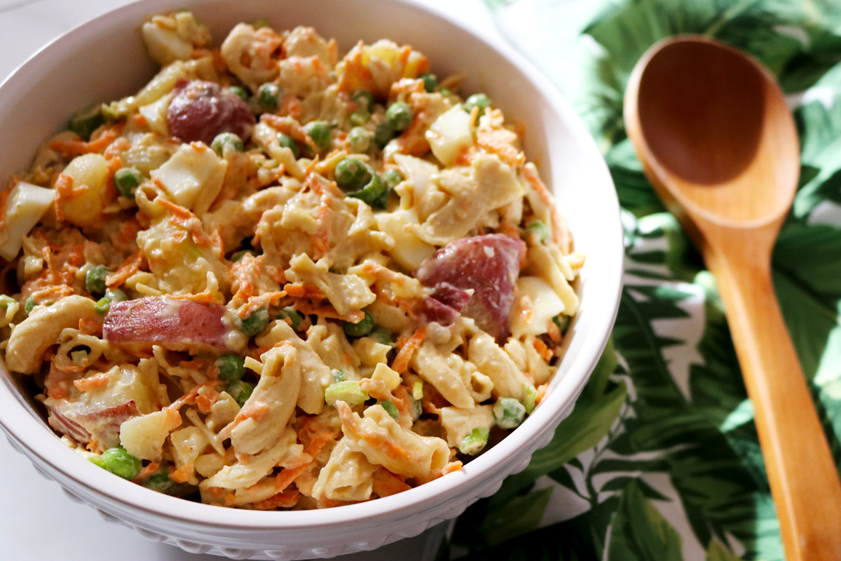 a bowl of Healthier Hawaiian-Style Potato-Mac Salad in a porcelain salad bowl next to a palm-print napkin and wooden serving spoon